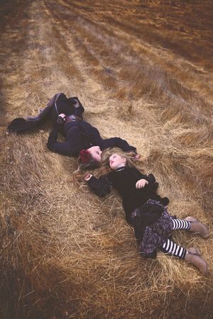 Two girls in dark gothic clothing and striped stockings lie head to head on dry golden straw in an overhead portrait.