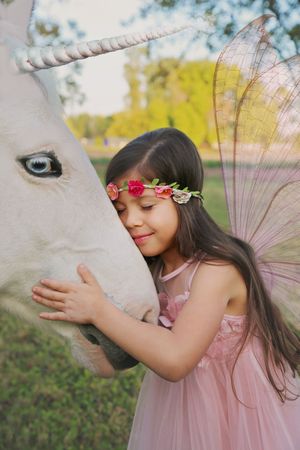 Girl in a pink fairy dress and flower crown gently hugs the face of a white unicorn with a spiral horn in a sunlit meadow.