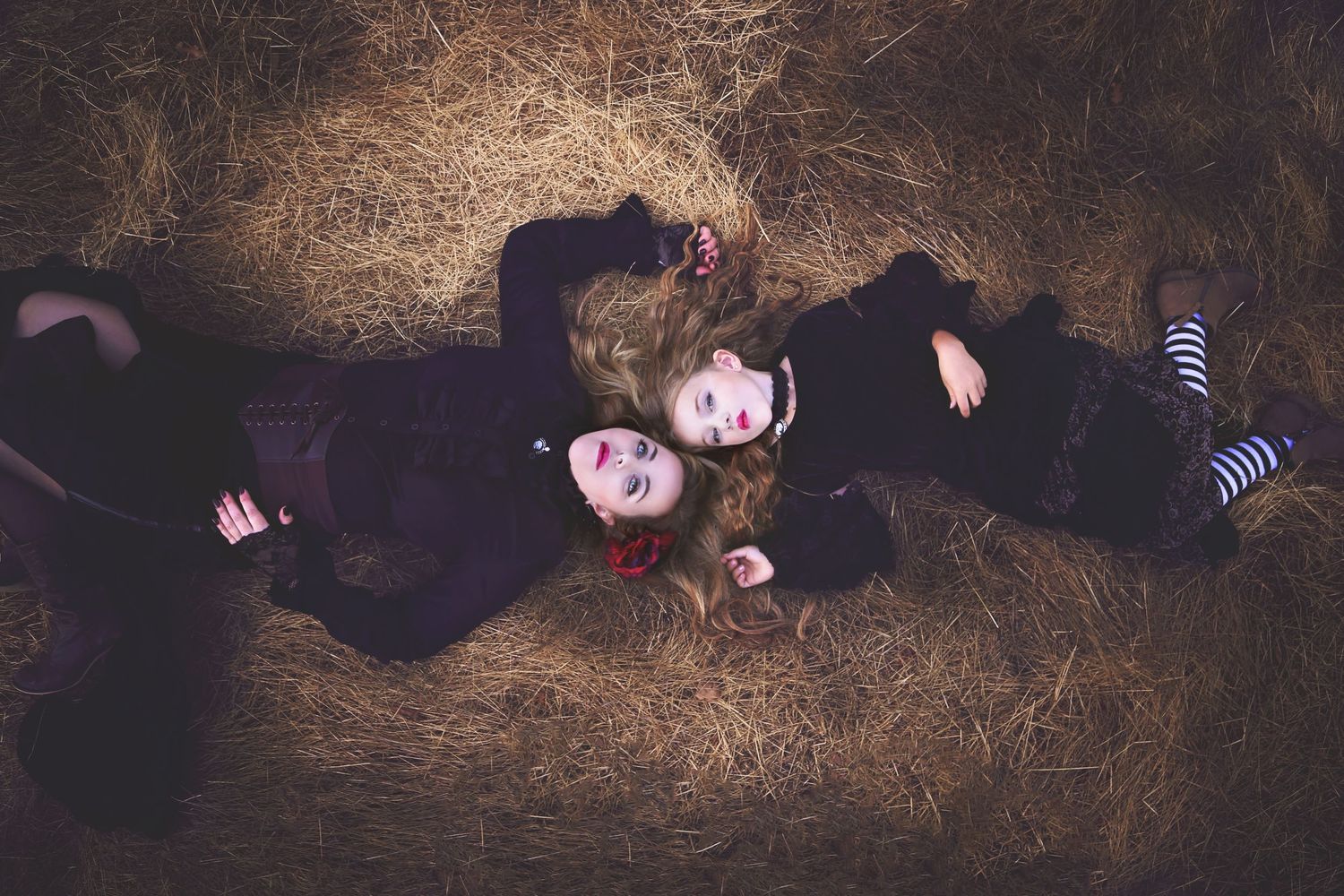 Overhead view of two girls in black gothic outfits lying on golden hay, one with a red flower in her hair.