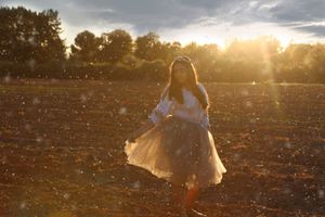 Girl in a flowing skirt twirls in a plowed field as golden sunset light and drifting particles fill the air.