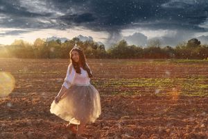 Woman in a flowing tulle skirt walks through a plowed field at golden hour beneath a dramatic stormy sky.