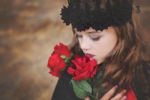 Young girl in a black lace crown holds red roses against her face in a moody, artistic fantasy portrait.