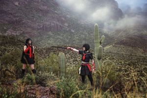 Two cosplayers in black and red costumes aim weapons at each other among cacti in a misty desert landscape.