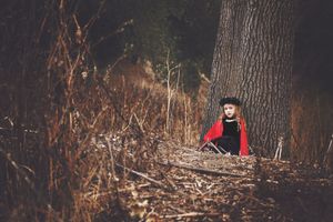 Small child in a red cape and black hat sits beside a large tree trunk in a dry, wintry autumn forest.