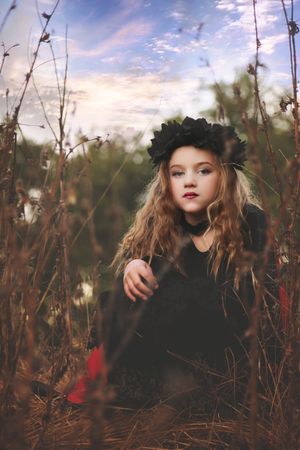 Girl with long wavy hair and a black flower crown sits among tall dry grass under a soft pastel sky.