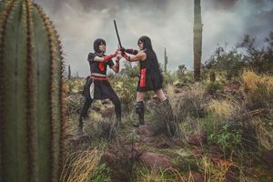 Two cosplayers in black and red costumes face off with swords in a desert landscape with tall saguaro cacti and dry grass.