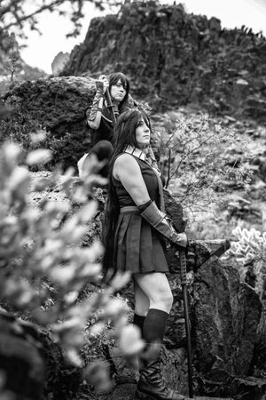 Black and white photo of two cosplayers in warrior costumes with gauntlets and boots posing among rocks and desert brush.