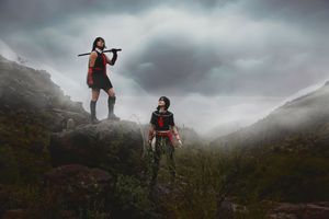 Two costumed cosplayers in dark red and black outfits, one holding a staff, stand on rocky mountain terrain beneath a moody, cloudy sky.