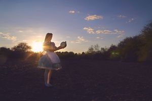 Silhouette of a woman in a light dress holding a small object in a dark field as the sun sets behind her.