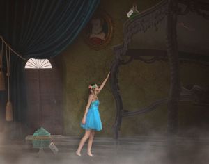 A barefoot ballerina in a turquoise dress reaches upward in a moody, vintage room with teal curtains, a portrait and a four-poster bed.