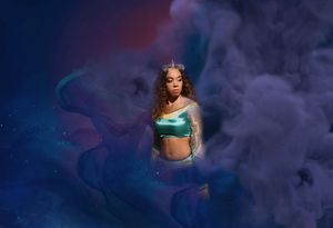 A woman in a teal satin top and a small crown stands amid swirling purple smoke against a dreamy blue cosmic backdrop.