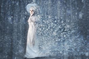 Ethereal woman in a white gown and headdress stands in a swirling blue snowstorm in a frozen forest fantasy scene.