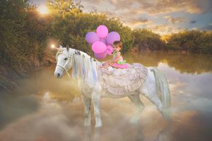 Child in a pink dress holding pink balloons rides a white horse standing in a reflective lake at sunset.