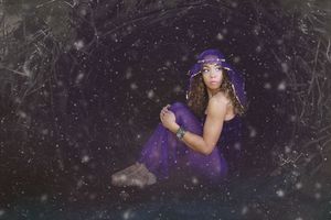 Woman in a purple veil and flowing gown sits curled up in a dark, mystical forest with sparkling lights floating around her.