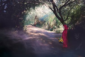 Sunlit dirt path winds through a leafy green forest with a woman in a red dress standing beside a tree holding yellow flowers.