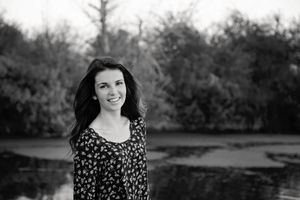 Black and white portrait of a smiling young woman with long dark hair in a floral top standing near a pond and trees.