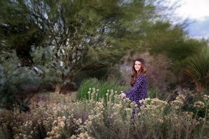 Woman in a dark patterned dress sits among blooming desert shrubs and green trees during a golden hour outdoor portrait session.