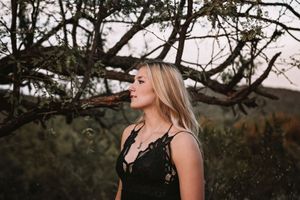 Blonde woman in a black lace top gazes into the distance beside a twisted mesquite tree branch in a warm, softly lit desert setting.