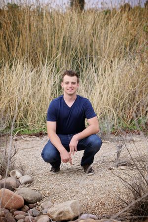 Smiling young man in a navy t-shirt and jeans crouches on a gravel path beside river rocks with tall golden grass behind him.