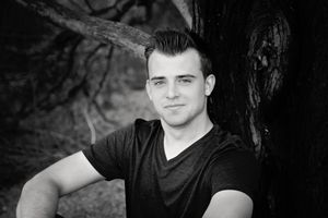 Black and white portrait of a young man in a dark t-shirt sitting beside a large tree trunk.