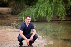 Young man in a navy shirt crouches on a sandy riverbank beneath green willow branches.