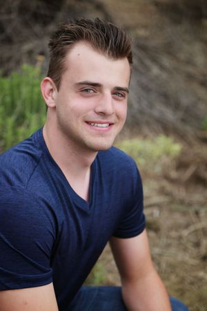 Smiling young man in a navy blue t-shirt poses for a close-up outdoor portrait with a natural green backdrop.