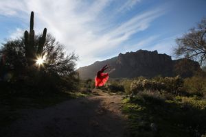 Dancer in a flowing red dress leaps on a desert trail near a saguaro cactus as the sun sets behind a rocky mountain.