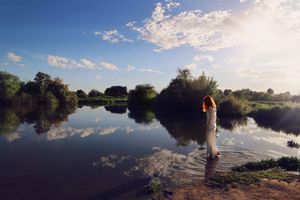 Woman in a white dress stands in shallow lake water at sunset, with soft clouds and trees reflected on the surface.