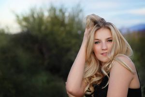 Portrait of a young woman with long wavy blonde hair running her hand through it against a blurred green desert background.