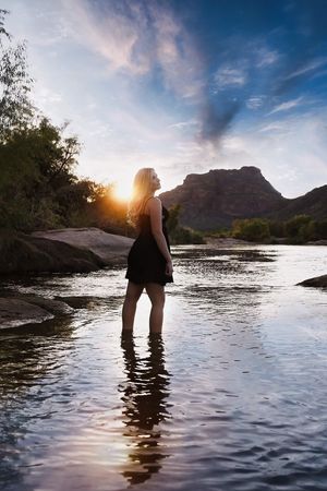 Woman in a black dress stands in shallow river water looking toward the setting sun, with a rocky mountain and colorful sky behind her.