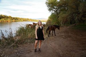 Young woman in a black dress standing on a dirt path beside a calm river with two horses grazing behind her in the soft evening light.