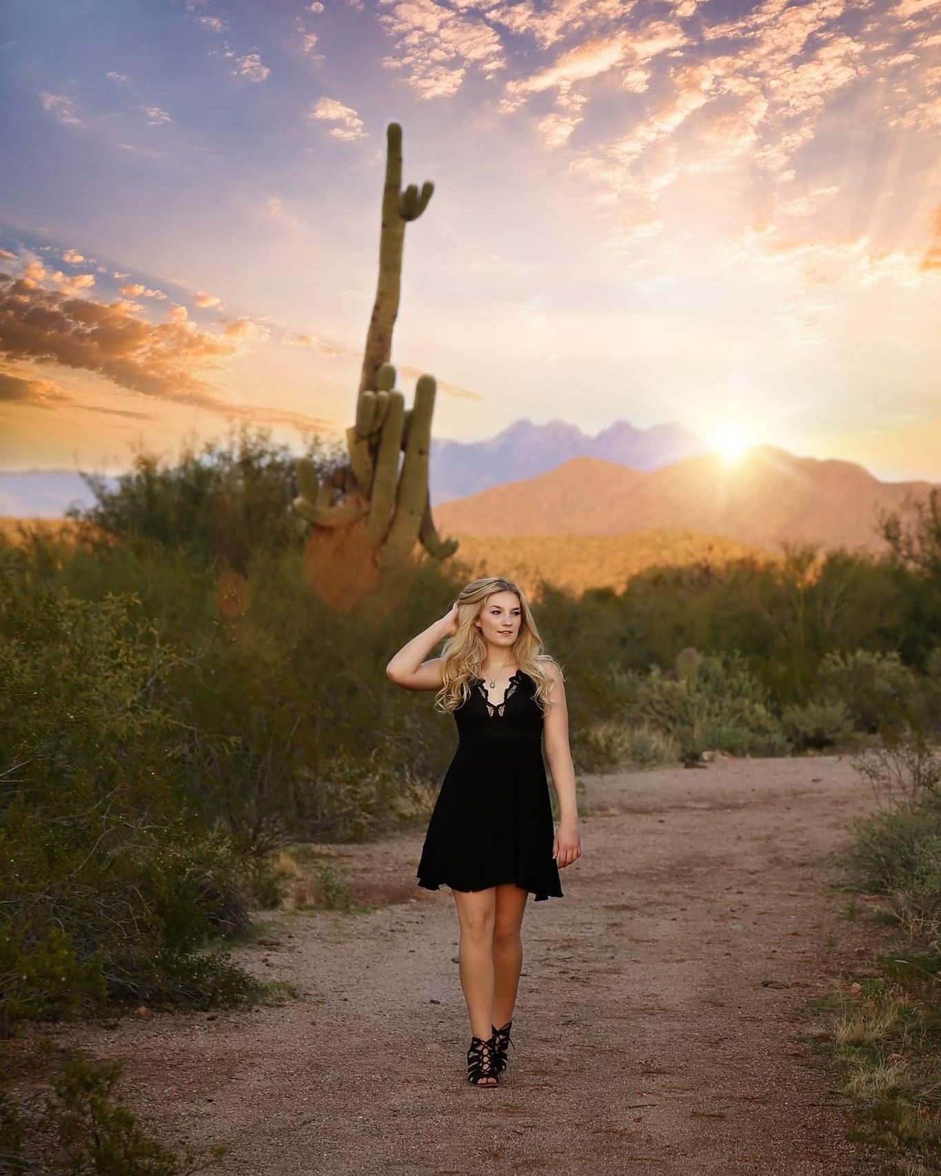 Young woman in a black dress walks along a desert trail at sunset near a tall saguaro cactus with mountains behind.