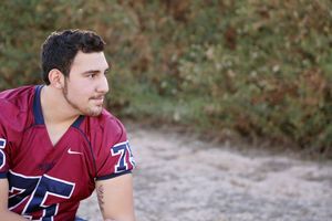 Young man with short dark hair wearing a maroon number 75 football jersey looks off to the side in front of green and autumn foliage.