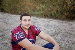 Smiling young man in a maroon number 75 football jersey sits on cracked dry earth in front of autumn-colored bushes for a senior portrait.