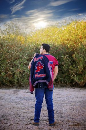 Young man in jeans and a maroon jersey stands facing golden bushes with a Pumas varsity jacket slung over his shoulder under a sunlit sky.