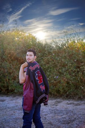 Young man looks over his shoulder while carrying a Pumas varsity letterman jacket, standing before golden bushes and a glowing sunset sky.