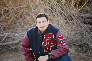 Young man in a navy and maroon Pumas letterman jacket with PP letters crouches outdoors in front of dry brush for a senior portrait.