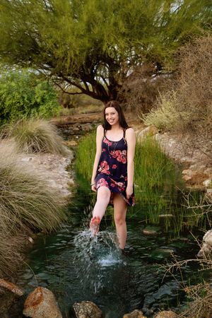 Smiling young woman in a navy floral dress splashes her foot in a clear desert stream surrounded by rocks, grass and a green tree.