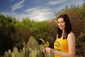 Smiling young woman in a yellow tank top holds small yellow wildflowers beside a prickly pear cactus under a blue sky.