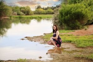 Young woman in a floral dress crouches at the edge of a calm pond with green grass and trees in warm evening light.