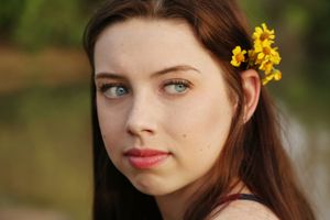 Close-up portrait of a young woman with long auburn hair and yellow flowers tucked behind her ear, glancing to the side.