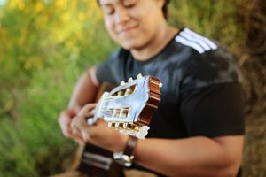 Young man in a black athletic shirt plays a classical guitar outdoors, with the guitar headstock in sharp focus against green foliage.