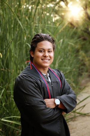 Smiling graduate in a black gown and red and blue honor cords stands with arms crossed in front of tall green grass and bright sunlight.