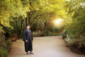 Graduate in a black cap-less gown stands on a tree-lined dirt path as golden sunlight and a faint rainbow flare glow behind him.
