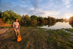 Man holds an acoustic guitar while standing on a grassy lakeshore at sunset, with trees and a soft blue sky behind him.