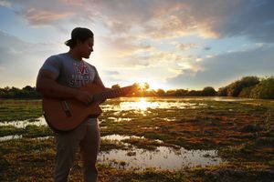 Man in a baseball cap plays an acoustic guitar beside a wetland at sunset, with golden light reflecting on the water.