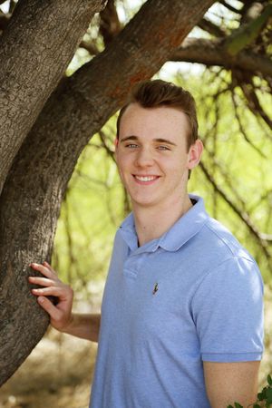 A smiling young man in a light blue polo shirt poses outdoors beside a tree trunk with soft green foliage behind him.