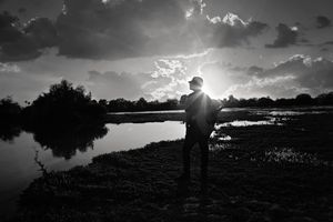 Black and white silhouette of a person standing beside a calm lake as the sun breaks through dramatic clouds.