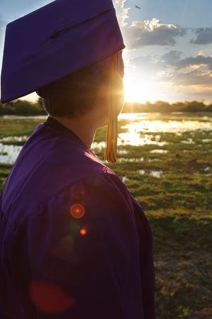 A graduate in a purple cap and gown with a gold tassel looks toward a glowing sunset over a wetland landscape.