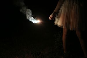 A woman in a tulle skirt holds a sparkler that glows and trails smoke against a dark night background.